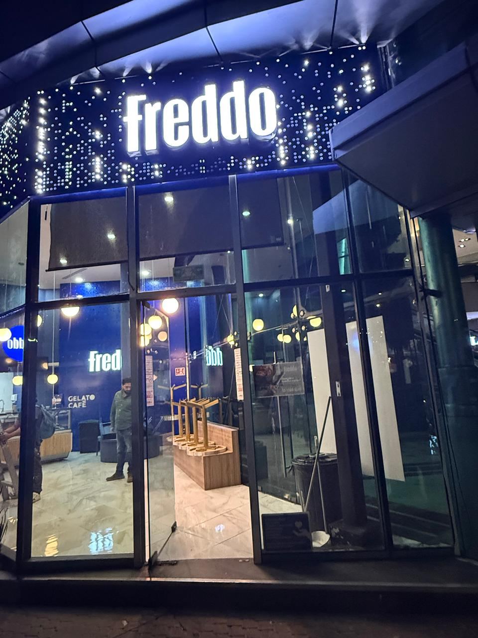 Fachada nocturna de la heladería Freddo con letrero luminoso sobre un tablero perforado retroiluminado