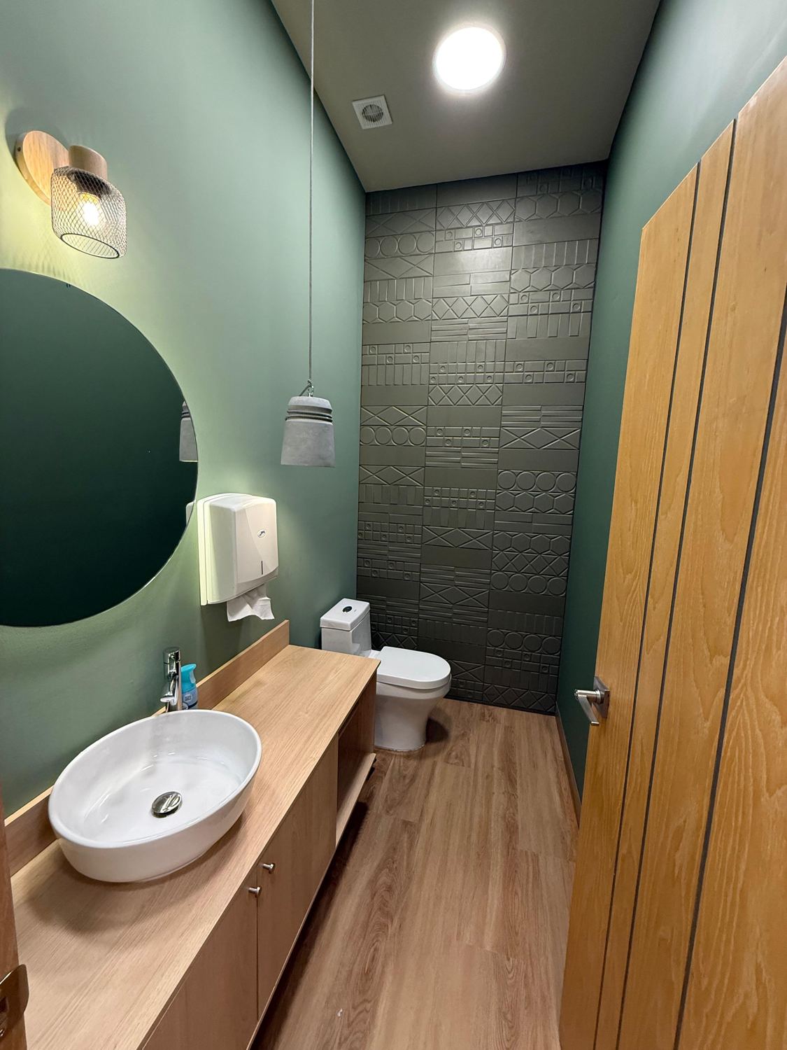 Baño con muro verde, muro con relieve geométrico, lavabo de sobreponer y puerta de madera